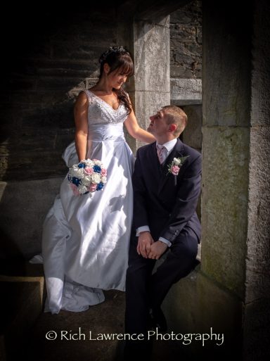 Bride in a white gown holds bouquet while looking at groom seated in formal attire.