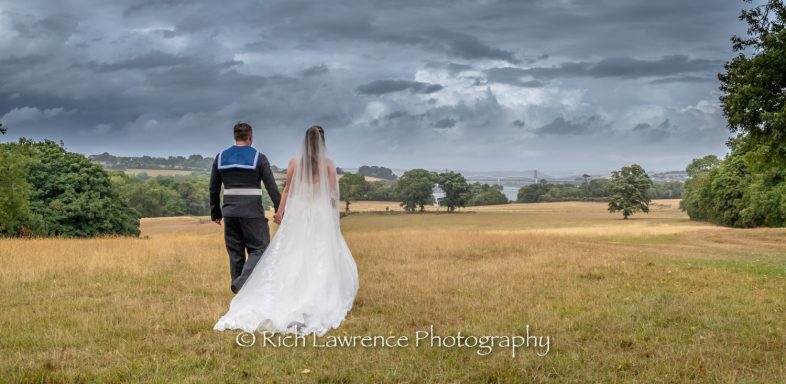 A couple in formal attire walking hand in hand across a grassy field under cloudy skies.