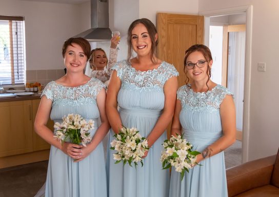 Four women in light blue dresses pose together, holding bouquets, smiling happily.