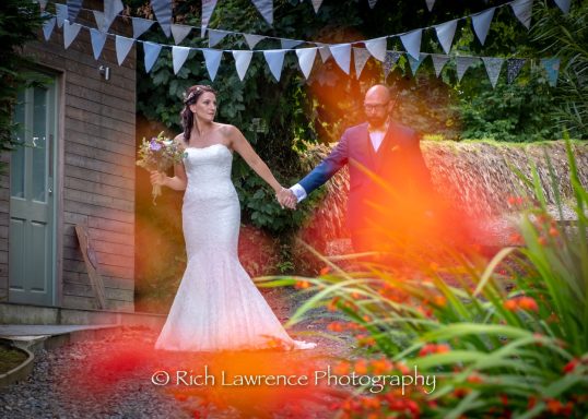 Bride and groom holding hands in a garden setting, surrounded by bunting and flowers.