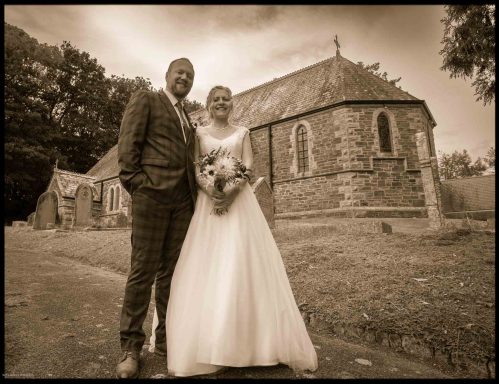 Bride and groom smiling together outside a stone church in a sepia-toned setting.