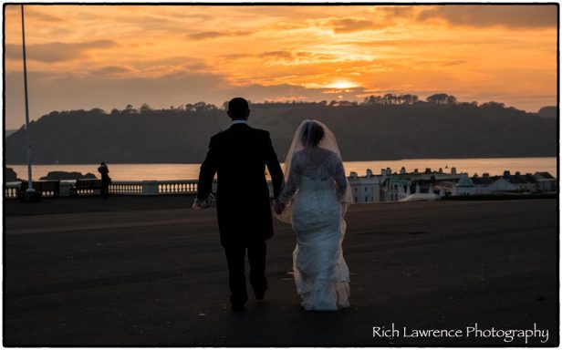 Bride and groom walking hand in hand at sunset by the water.