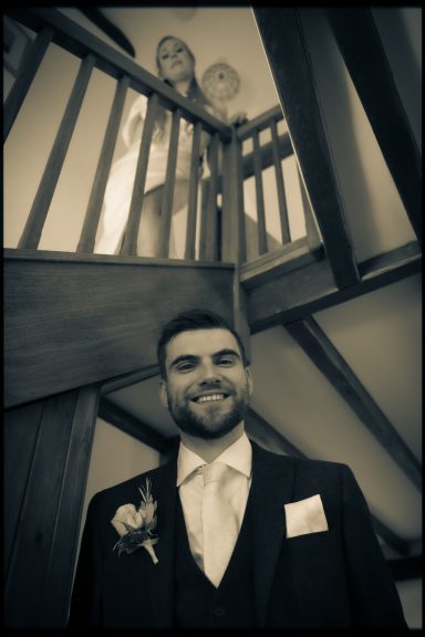 Groom smiling at the camera, with two guests peering from the staircase above.