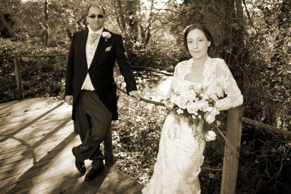 A bride and groom pose together on a bridge in a forest setting.