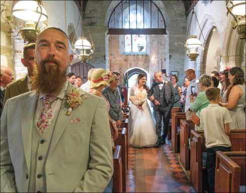A bearded man in a suit stands in a wedding ceremony with guests and the bride entering.