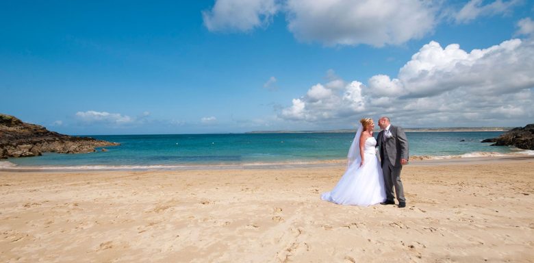 Bride and groom standing on a sandy beach with a blue sea and sky in the background.