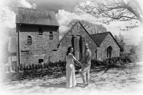 A couple holds hands in front of a rustic stone church with trees in the background.