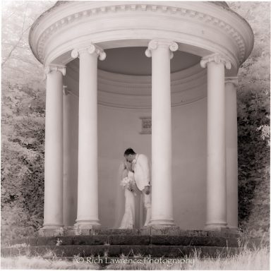 A couple in formal attire standing in a white, columned gazebo surrounded by greenery.