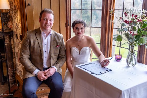 A groom in a tan suit and a bride in a strapless gown seated at a table with flowers.
