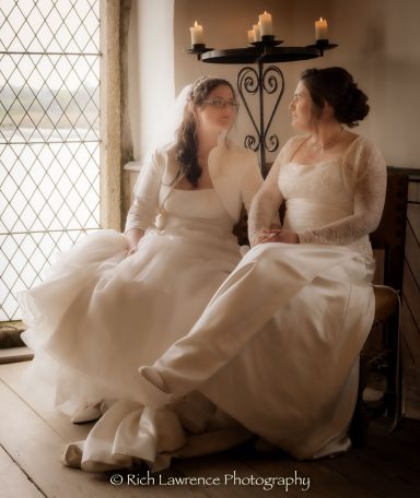 Two brides in elegant wedding dresses sit together by a window, smiling softly at each other.