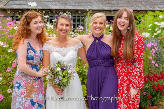 Four women smiling together in a garden, one in a wedding dress, surrounded by flowers.
