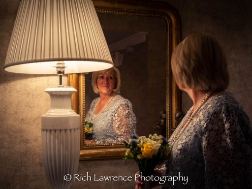 A woman in a silver dress holding flowers, reflected in a mirror beside a lamp.