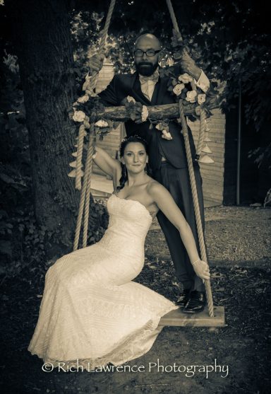 A bride in a white gown and a groom in a suit pose together on a swing in a wooded setting.