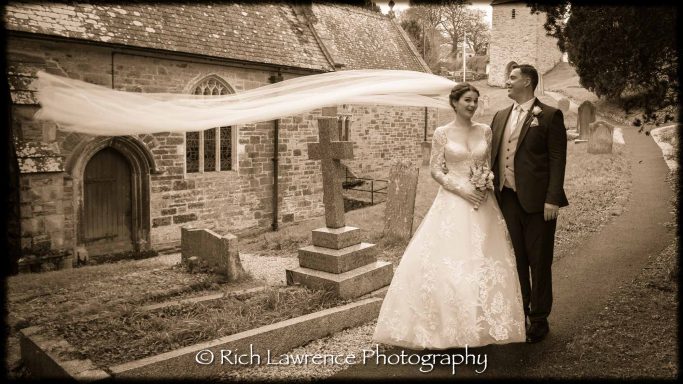 Bride and groom standing outside a church, with a flowing veil and a gravestone nearby.