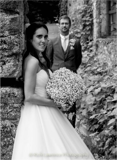 A bride in a strapless gown holds a bouquet, with a groom smiling in the background.