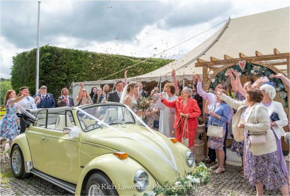 A joyful wedding scene with guests celebrating near a vintage yellow car.