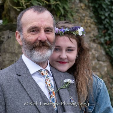A smiling man in a grey suit stands behind a young woman with flowers in her hair.