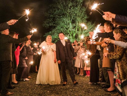 A newlywed couple surrounded by guests holding sparklers at night.