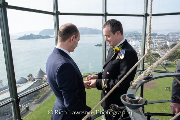 Two men exchanging rings with a coastal view in the background.