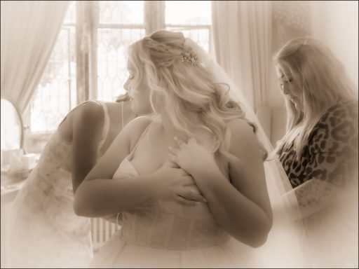 A bride adjusting her dress with two other women assisting in a softly lit room.