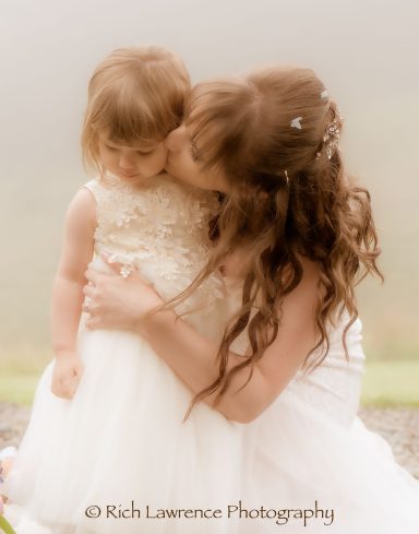 A bride lovingly kisses a young girl in a white dress against a soft, misty background.
