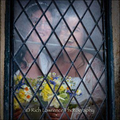 Two smiling women in wedding dresses, holding a bouquet, seen through a diamond-patterned window.