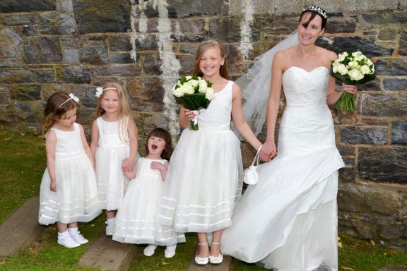 A bride in a white gown stands with four young girls in white dresses, smiling and holding flowers.