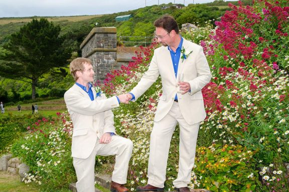 Two well-dressed men in light suits shake hands among vibrant flowers in a garden.