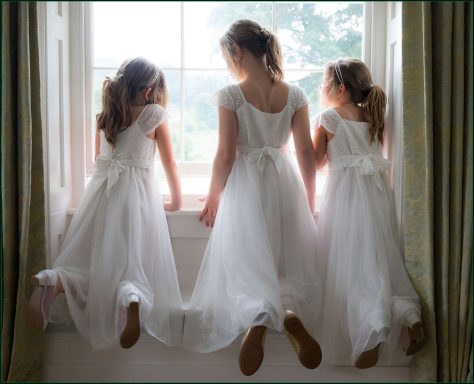 Three young girls in white dresses sit on a windowsill, gazing outside.