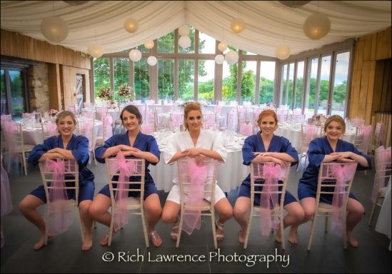 Five women in blue robes seated on chairs with pink sashes in a decorated venue.