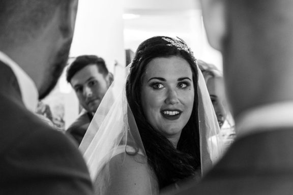 Bride smiling warmly, surrounded by guests, wearing a veil in soft lighting.