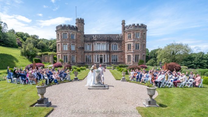 A wedding ceremony held in front of a large castle, surrounded by guests and greenery.