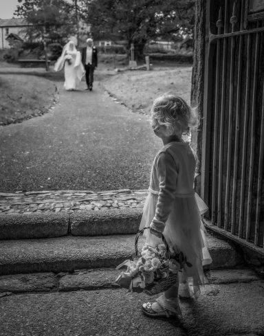 A young girl holding flowers watches a bride and groom walking down a path.